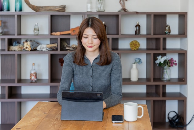 A young woman smiling while using a tablet at a wooden table with a mug and smartphone.