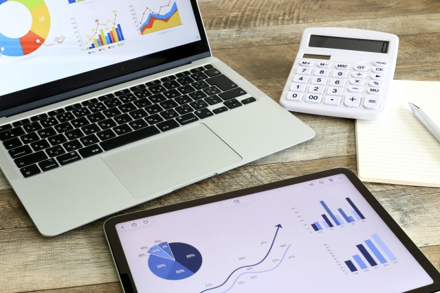 Laptop and tablet displaying financial charts and graphs next to a calculator and notepad on a wooden desk.