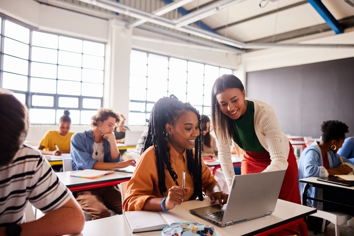 A smiling teacher leaning over a laptop to assist a student in a bright, modern classroom.