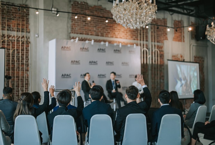 Audience members raise their hands during a Q&A session at an APAC panel discussion in an industrial-style venue.