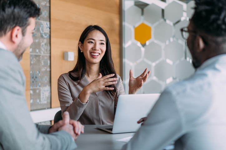 A woman smiling and gesturing during a business meeting with colleagues in a modern office.